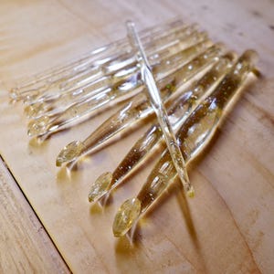 May include: A set of clear crochet hooks with gold glitter embedded in the plastic. The hooks are arranged in a fan shape on a wooden surface.