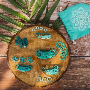 May include: A wooden round board with various sizes of teal and green plastic rings, a set of teal and green plastic hooks, and a teal drawstring bag with a white mandala design.