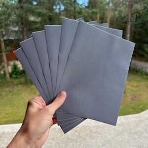 Könnte beinhalten: Eine Hand hält einen Stapel grauer Notizbücher. Die Notizbücher sind aufgefächert und zeigen ihre rechteckige Form und die Textur des Papiers. Der Hintergrund zeigt eine verschwommene Ansicht von grünen Bäumen.