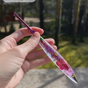 Puede incluir: Un ganchillo hecho a mano con un mango de resina transparente, incrustado con flores moradas y rojas y copos de oro. El gancho tiene un acabado púrpura metálico. La palabra "Woolpedia" está en la esquina superior izquierda.