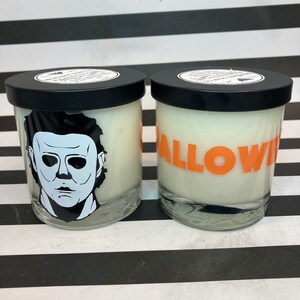 May include: Two glass candles with black lids. One candle features a black and white graphic of a face, and the other candle has the word "HALLOWEEN" in orange letters. The candles are on a black and white striped surface.