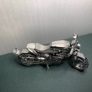 May include: A silver metal motorcycle model with a black seat and wheels. The motorcycle is on a green surface.