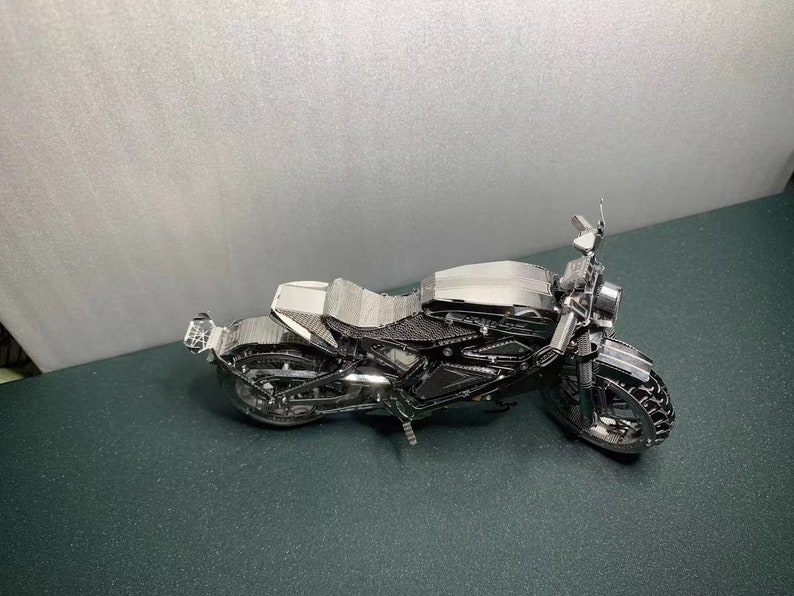 May include: A silver metal motorcycle model with a black seat and wheels. The motorcycle is on a green surface.
