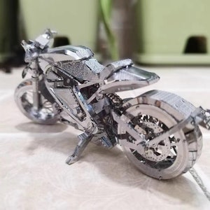 May include: A silver metal model motorcycle with intricate details. The motorcycle has a black seat and a silver exhaust pipe.