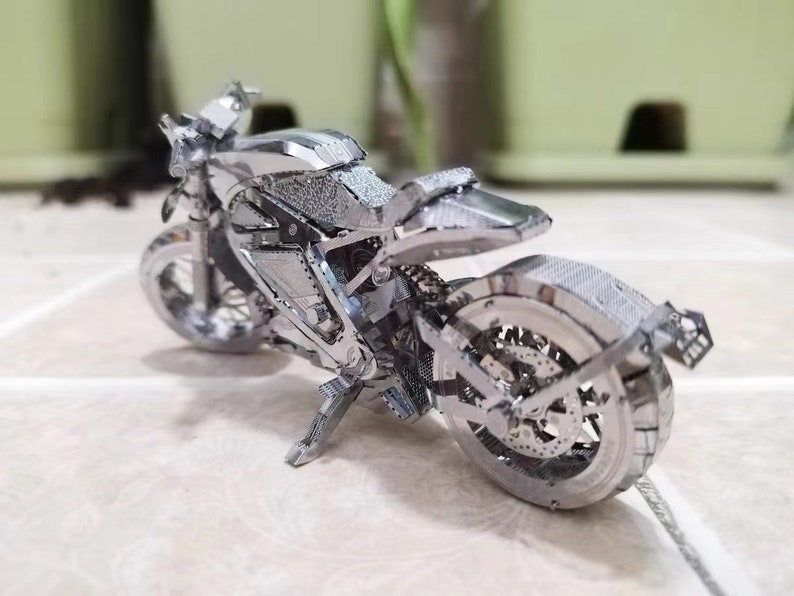 May include: A silver metal model motorcycle with intricate details. The motorcycle has a black seat and a silver exhaust pipe.