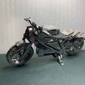 May include: A silver metal model motorcycle with a black seat and wheels. The motorcycle is detailed with a chrome finish and has a kickstand.
