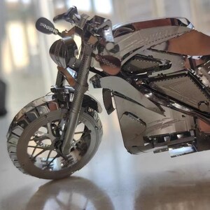 May include: A silver metal model motorcycle with a detailed design. The motorcycle has a front wheel, handlebars, and a seat.