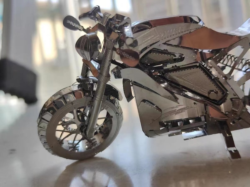 May include: A silver metal model motorcycle with a detailed design. The motorcycle has a front wheel, handlebars, and a seat.