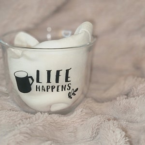 Peut inclure: Une tasse en verre transparent à double paroi. La tasse a une poignée noire et un motif noir qui dit "Life Happens" avec un petit motif de feuille.