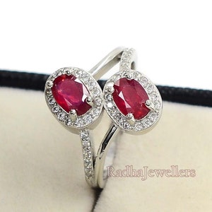 Puede incluir: Un anillo bypass de tono plateado con dos rubíes rojos de corte ovalado, cada uno rodeado por un halo de pequeñas piedras claras. La banda está dividida y acentuada con pequeñas piedras claras. El texto RadhaJewellers es visible en el anillo.