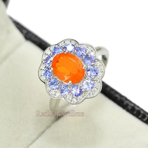 May include: A silver ring with an orange oval gemstone surrounded by smaller blue gemstones and white diamonds. The ring is in a black ring box.