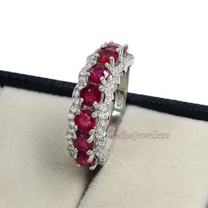 Puede incluir: Un anillo plateado con una fila de gemas rojas vibrantes, intercaladas con piedras claras más pequeñas. El anillo se presenta en una caja de joyería negra con interior blanco. Primer plano de los detalles de las gemas.