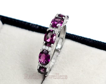 May include: A silver ring featuring a row of oval-cut, deep purple gemstones. Small, clear accent stones are interspersed between the larger gems. The ring is displayed in a black jewelry box with a white interior.
