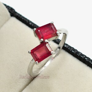 Könnte beinhalten: Ein silberner Ring mit zwei rechteckigen, leuchtend roten Edelsteinen. Der Ring wird in einer schwarzen Schmuckschatulle mit hellem Innenraum präsentiert, was den Kontrast hervorhebt. Die roten Edelsteine sind der Fokus.