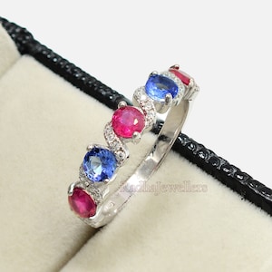 May include: A silver band ring with alternating red and blue gemstones. The ring is set in a black velvet ring box.