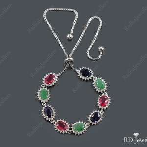 May include: A silver-toned adjustable bolo bracelet featuring a series of oval-cut red, green, and dark blue gemstones. Each stone is surrounded by a halo of small white crystals. The bracelet has a box chain with two dangling metal beads.