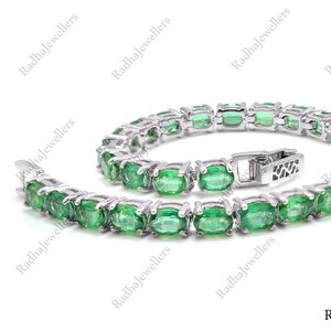 Puede incluir: Una pulsera de tenis en tono plateado que presenta una hilera continua de piedras preciosas ovaladas de color verde vibrante, engastadas en garras metálicas individuales. La pulsera incluye un cierre de caja seguro y está dispuesta en círculo.