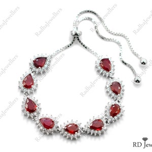 May include: A silver-toned adjustable bolo bracelet featuring seven pear-shaped deep red gemstones, each surrounded by a halo of small clear stones. The bracelet has a delicate box chain with two dangling ends and a sliding clasp mechanism.