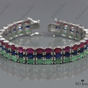 Könnte beinhalten: Silberarmband mit drei Reihen farbiger Edelsteine. Die oberen Reihen zeigen rote, blaue und grüne Steine. Das Armband ist geöffnet und zeigt den Verschlussmechanismus. Ein elegantes Schmuckstück.