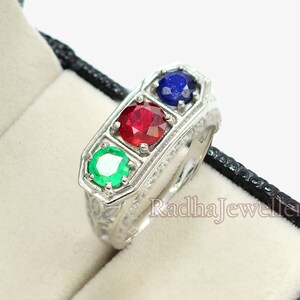 May include: A silver ring with three gemstones: a green emerald, a red ruby, and a blue sapphire. The ring is set in a decorative, engraved band and is displayed in a black jewelry box with a cream-colored interior.