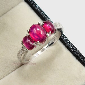 Peut inclure: Une bague en argent ornée de trois pierres précieuses ovales rouge vif. La bague est sertie dans un anneau décoratif avec des détails gravés. La bague est présentée dans un écrin noir avec un intérieur beige clair.