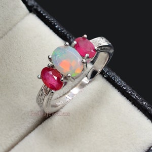 May include: A silver ring featuring an oval opal gemstone flanked by two oval ruby gemstones. The opal has a play of color, and the rubies are a deep red. The ring is set in a silver band with decorative detailing.