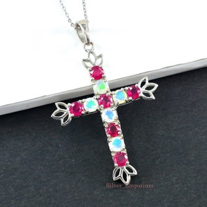 May include: A silver cross pendant necklace with a delicate chain. The cross is adorned with alternating red and iridescent gemstones. The pendant has decorative floral accents at the ends of the cross arms and at the top.