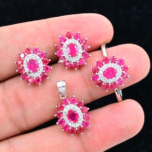 May include: A jewelry set featuring a ring, pendant, and earrings. Each piece has a central oval ruby surrounded by smaller rubies and a halo of white gemstones. The set is displayed against a black background.
