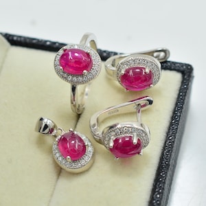 Puede incluir: Un conjunto de joyería que incluye un anillo, un par de pendientes y un colgante. Cada pieza presenta una piedra central de color rosa vibrante con corte cabujón ovalado, rodeada por un halo de pequeñas piedras claras en un marco plateado.
