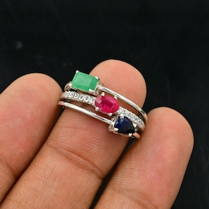 Puede incluir: Un anillo de plata con tres piedras preciosas: una esmeralda verde, un rubí rojo y un zafiro azul. El anillo está engastado con pequeños diamantes.