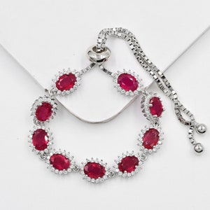 May include: A silver-toned adjustable bolo bracelet featuring eight oval-cut deep red stones, each surrounded by a halo of small clear white stones. The bracelet has a delicate box chain with a sliding clasp mechanism and two small metal beads.