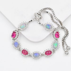 May include: A silver-toned adjustable bracelet featuring alternating oval-shaped iridescent opals and vibrant pink stones. Each stone is surrounded by a halo of small clear crystals, connected by a box chain with a sliding clasp mechanism.