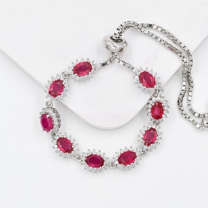 May include: A silver-toned adjustable bracelet featuring a series of oval-cut vibrant red stones, each surrounded by a halo of small clear white crystals. The bracelet includes a sliding bead mechanism and a delicate box chain for a secure fit.