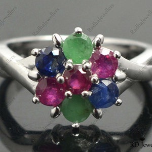 May include: A silver ring featuring a cluster of gemstones in the centre. The gemstones are red, green, and blue. The ring is set with a white metal band.