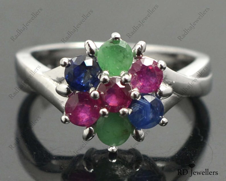 May include: A silver ring featuring a cluster of gemstones in the centre. The gemstones are red, green, and blue. The ring is set with a white metal band.