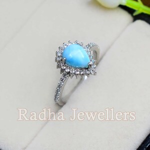 May include: A pear-shaped turquoise gemstone ring with a halo of small clear stones. The ring is set in silver and has a delicate band.