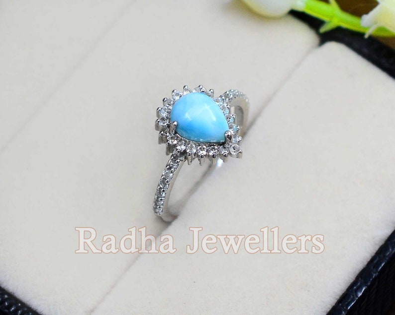 May include: A pear-shaped turquoise gemstone ring with a halo of small clear stones. The ring is set in silver and has a delicate band.