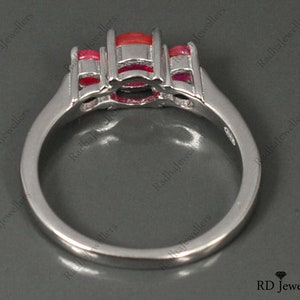 May include: A silver ring with a large red gemstone and smaller red gemstones on the sides. The ring is set with a white metal band.
