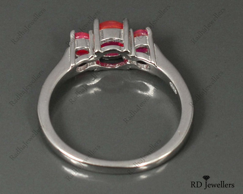 May include: A silver ring with a large red gemstone and smaller red gemstones on the sides. The ring is set with a white metal band.