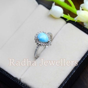 May include: A pear-shaped turquoise ring with a halo of small diamonds set in silver. The ring is displayed in a black ring box with white velvet lining. The text "Radha Jewellers" is visible on the box.
