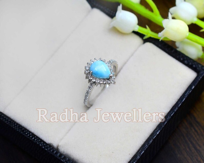 May include: A pear-shaped turquoise ring with a halo of small diamonds set in silver. The ring is displayed in a black ring box with white velvet lining. The text "Radha Jewellers" is visible on the box.