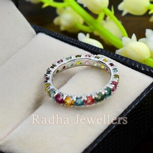 May include: A white gold eternity band ring with a row of colorful gemstones. The ring is set in a white gold band and features a variety of gemstones, including pink, green, blue, yellow, and orange. The ring is displayed in a black velvet ring box.