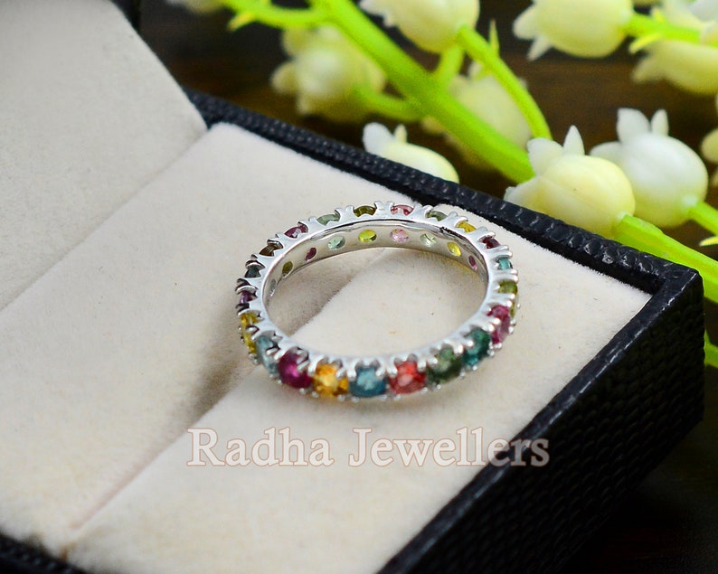 May include: A white gold eternity band ring with a row of colorful gemstones. The ring is set in a white gold band and features a variety of gemstones, including pink, green, blue, yellow, and orange. The ring is displayed in a black velvet ring box.