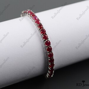 May include: A silver-toned tennis bracelet featuring a continuous row of round, deep red rubies set in individual metal prongs. The bracelet is curved over a white cylindrical form, showcasing the uniform size and vibrant color of each gemstone.