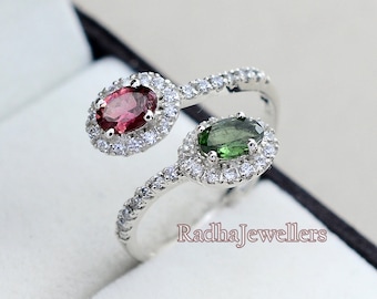Può includere: Un anello d'argento con due pietre ovali incastonate in un alone di diamanti più piccoli. L'anello presenta una pietra rosa e una pietra verde.