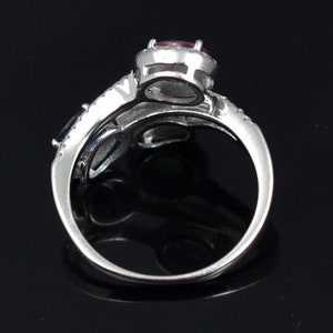 Op de afbeelding: Een zilveren ring met een grote, ronde, rode edelsteen in het midden, omringd door kleinere, heldere stenen. De band splitst zich in twee&euml;n, met een blauwe edelsteen aan &eacute;&eacute;n kant. De ring staat tegen een zwarte achtergrond.