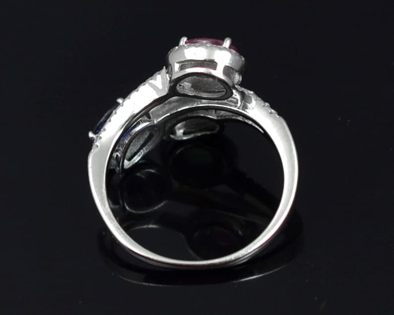 Op de afbeelding: Een zilveren ring met een grote, ronde, rode edelsteen in het midden, omringd door kleinere, heldere stenen. De band splitst zich in twee&euml;n, met een blauwe edelsteen aan &eacute;&eacute;n kant. De ring staat tegen een zwarte achtergrond.