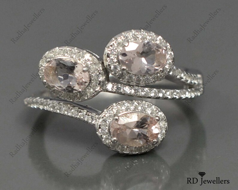 May include: A silver ring with three oval-shaped pink gemstones surrounded by smaller clear gemstones. The ring has a twisted band.