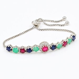 May include: A silver-toned adjustable bolo bracelet featuring a central circular charm set with a bright pink stone surrounded by small clear crystals, flanked by alternating round blue, green, and pink stones in prong settings on a box chain.