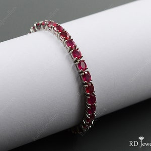 May include: A silver-toned tennis bracelet featuring a continuous row of round, vibrant red rubies set in metal prongs. The jewelry piece displays a uniform design with faceted gemstones that catch the light, creating a classic and elegant appearance.